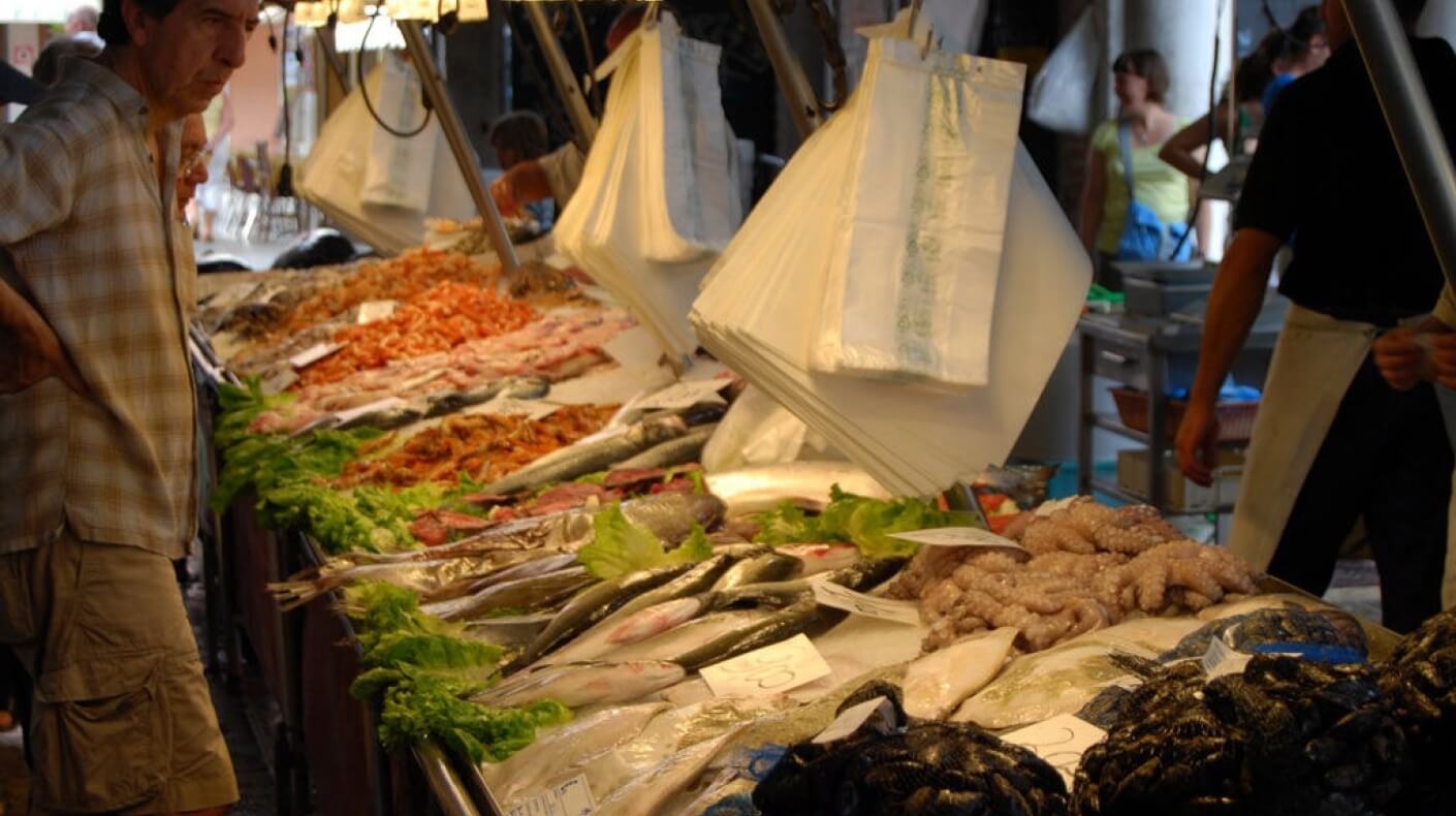 Un mercato affollato con un banco di frutti di mare, un uomo in camicia a quadri esamina le offerte mentre parla con un altro.