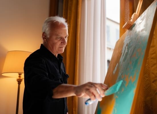 Uomo anziano con capelli bianchi dipinge su una grande tela gialla, con una finestra e tende gialle sullo sfondo.