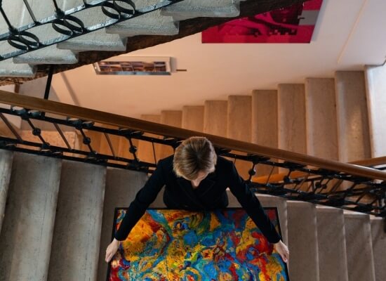 Una donna sale le scale con un grande dipinto astratto, vista da dietro, su gradini in cemento e corrimano in legno.