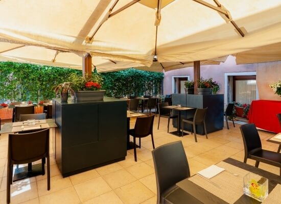 Patio di ristorante moderno con tetto bianco, tavoli apparecchiati e piante, circondato da una siepe verde.