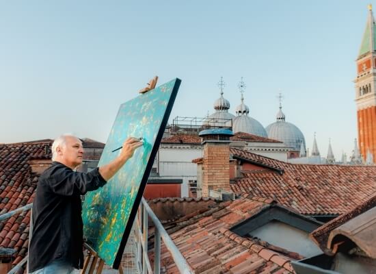 Uomo su un tetto dipinge un grande quadro astratto, con un panorama di tetti e un edificio bianco sullo sfondo.