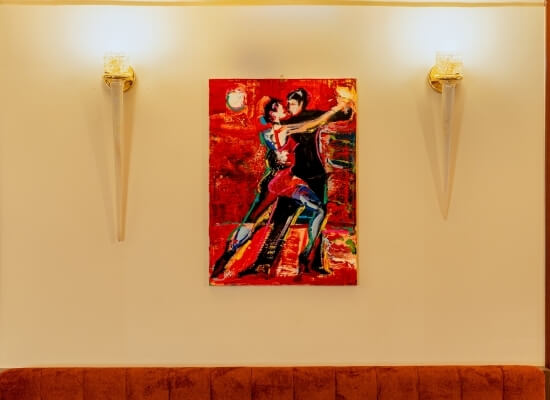 Coppia danzante in un dipinto rosso su muro beige, illuminato da due applique, con superficie marrone in basso.