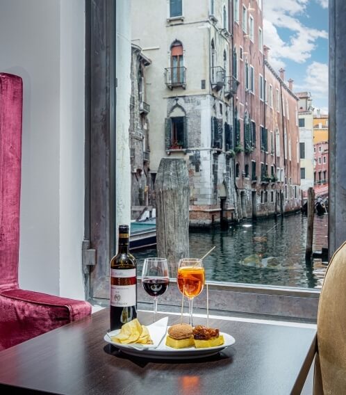 Tavolo in legno scuro con cibo e bevande, vista su un canale attraverso una grande finestra aperta.