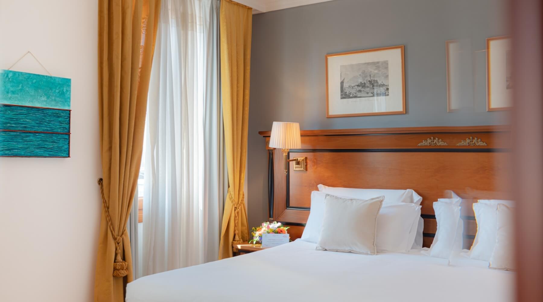 Camera da letto serena con letto bianco, testata in legno, lampada, fiori e luce naturale che filtra dalle tende gialle.