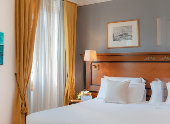 Hotel room featuring a king-sized bed with white linens, yellow curtains, and a nightstand with a lamp and fruit tray.