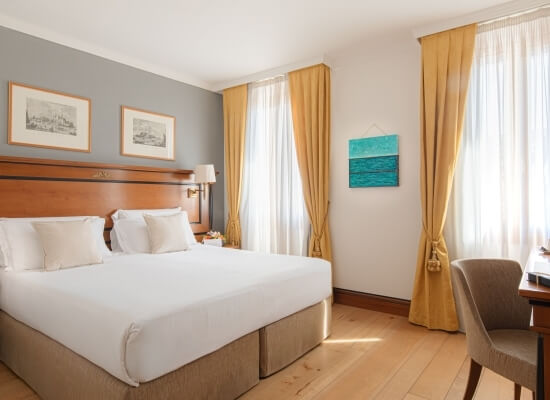 Hotel room with a double bed, two windows with yellow curtains, decorative elements, and natural light.