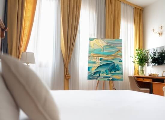 Una stanza serena con un dipinto astratto blu, giallo e bianco, un letto, una lampada e un desk con fiori bianchi.
