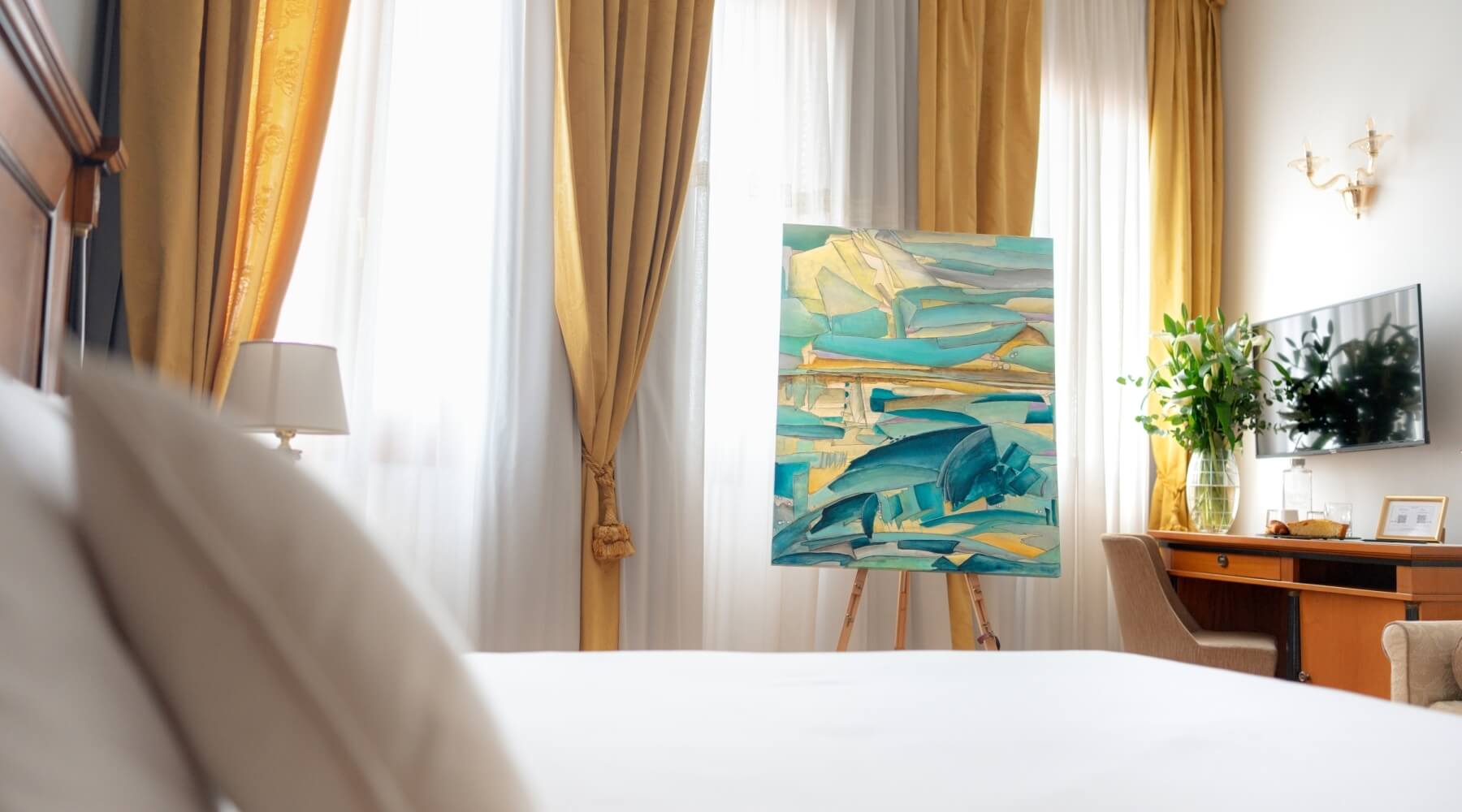 Una stanza con un grande dipinto su un cavalletto, letto bianco, lampada, vaso di fiori, TV e finestra con tende gialle.