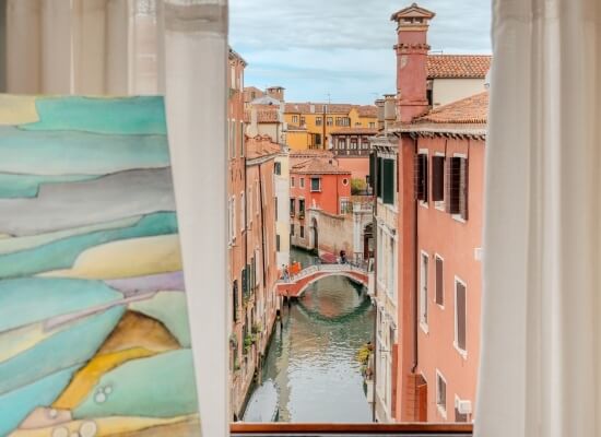 Finestra con tende bianche che incorniciano un canale di Venezia e un dipinto vibrante a sinistra, ricco di colori e forme.