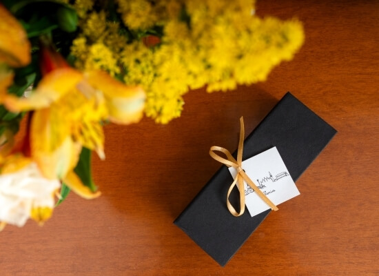Scatola regalo nera con nastro giallo e biglietto su un tavolo di legno, accanto a un bouquet di fiori gialli.