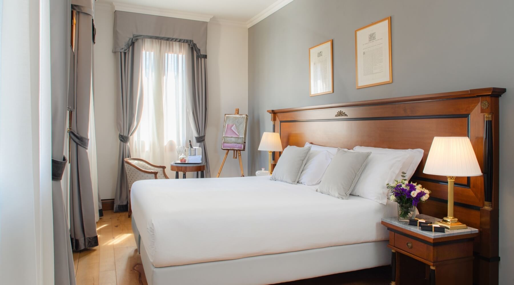 Camera serena con letto grande, biancheria bianca, comodino, lampada, fiori, sedia, cavalletto e luce naturale.