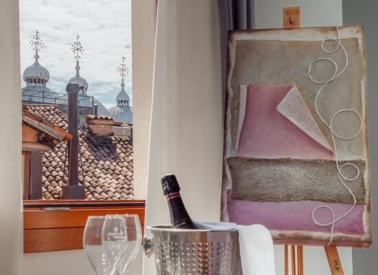 Interno artistico con finestra, dipinto astratto, champagne e vista su tetti sotto un cielo grigio.