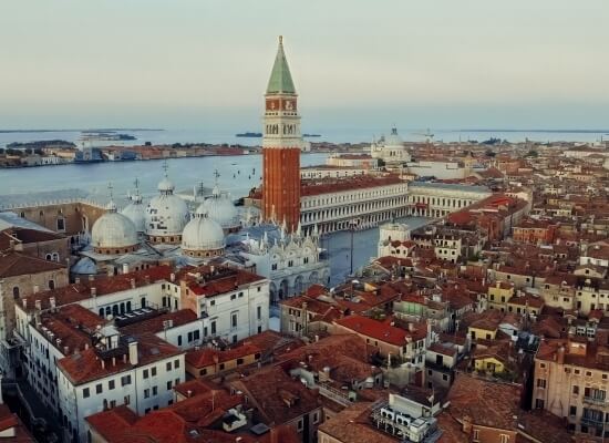Vista aerea di una città con un torre centrale, edifici bianchi e tetti rossi, sullo sfondo di acqua e cielo blu.