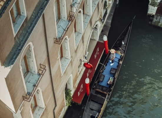 Vista aerea di una gondola nera ormeggiata vicino a un edificio tan, con un gondoliere e un passeggero in un cappello di paglia.