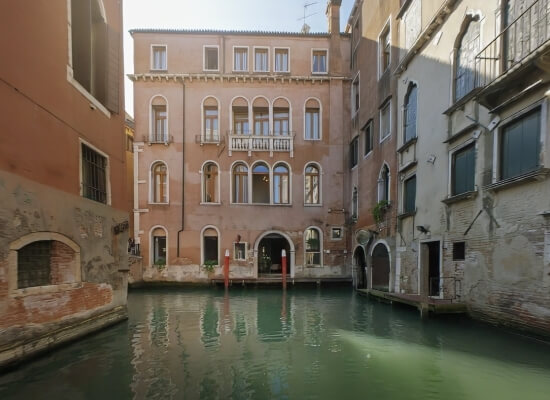 Canale sereno a Venezia con edifici color salmone e pali arancioni, riflessi nell'acqua verde-blu. Cielo azzurro.