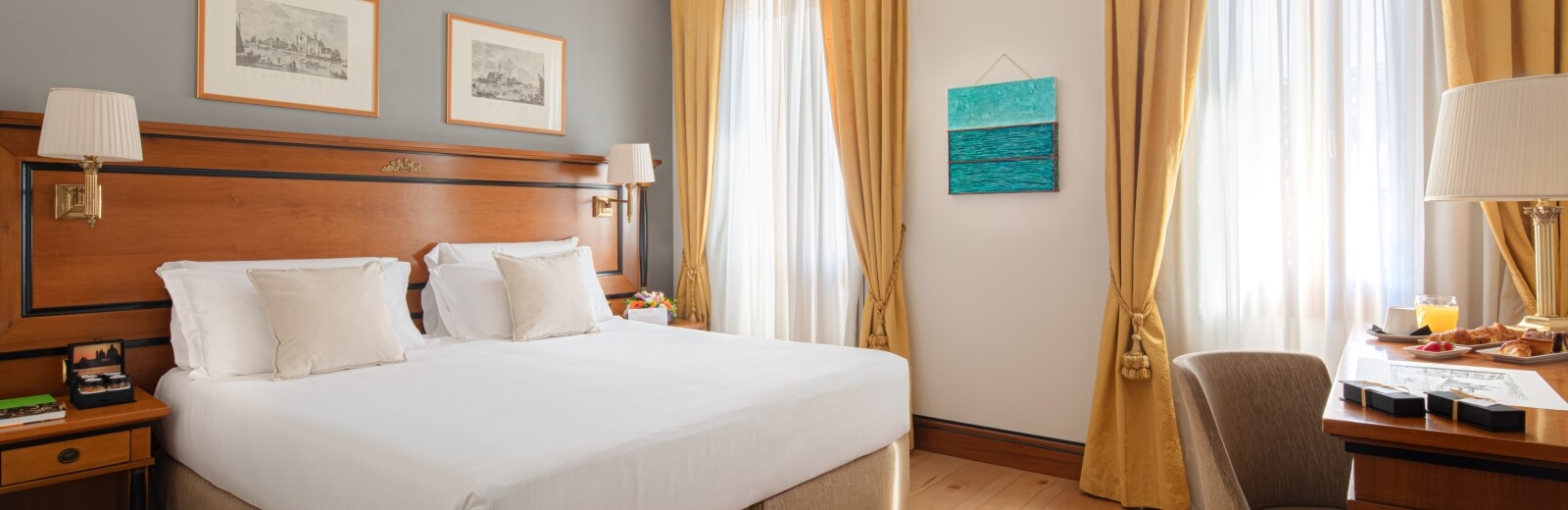 A cozy bedroom with a large bed, bedside lamps, a dining table with croissants, and sunlight streaming through a gold curtain.