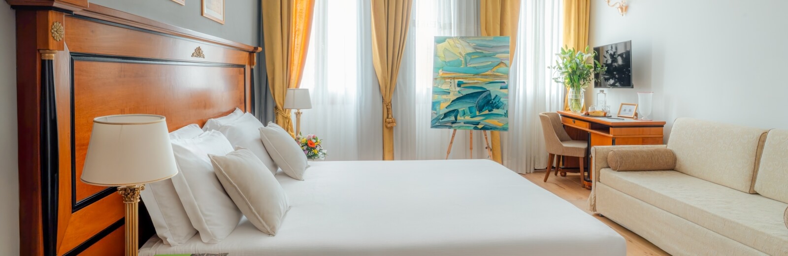 Camera d'albergo elegante con letto grande, divano bianco, scrivania e luce naturale che entra dalle finestre.