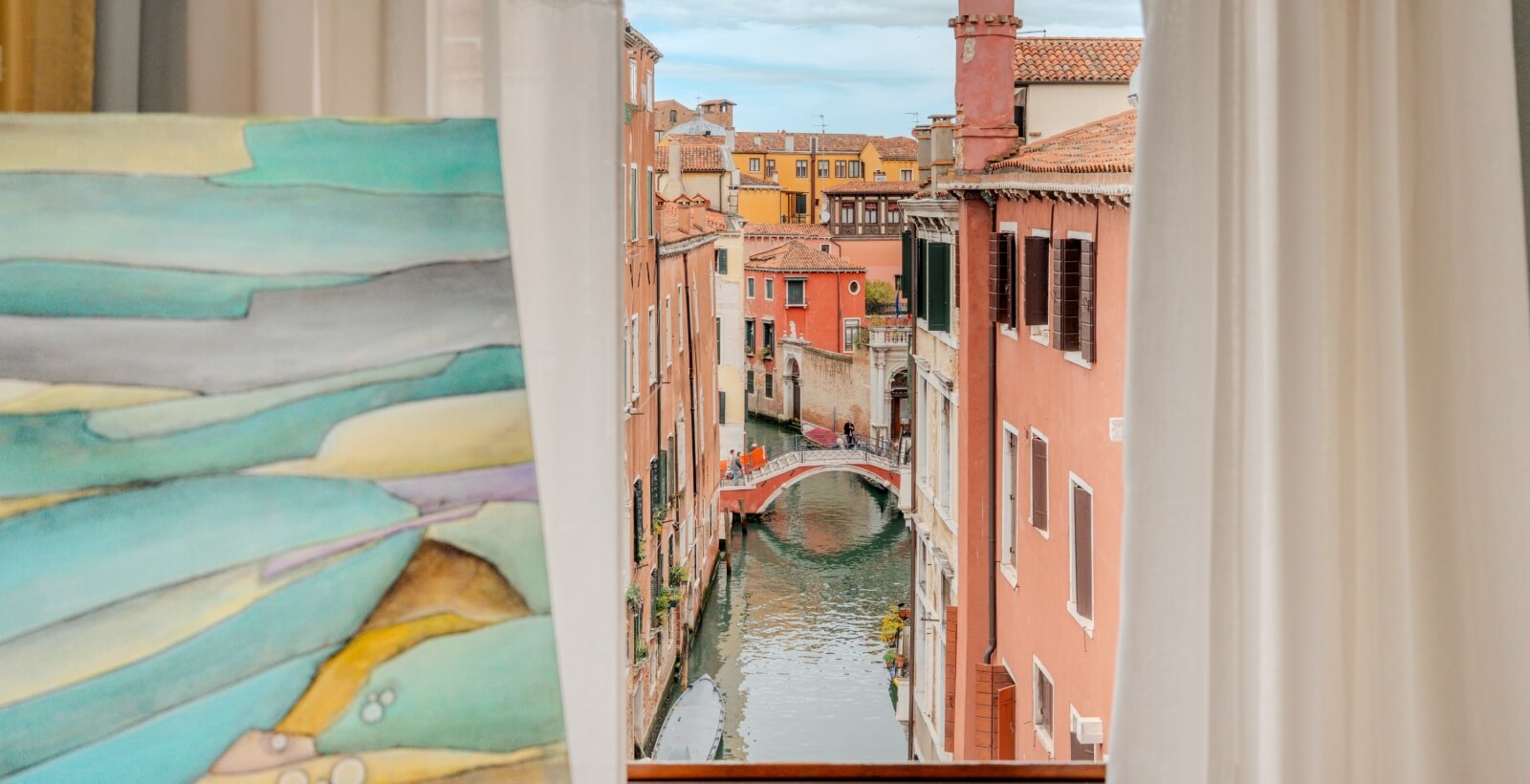 Vista serena di un canale di Venezia, incorniciato da tende bianche, con edifici colorati e un ponte ad arco.