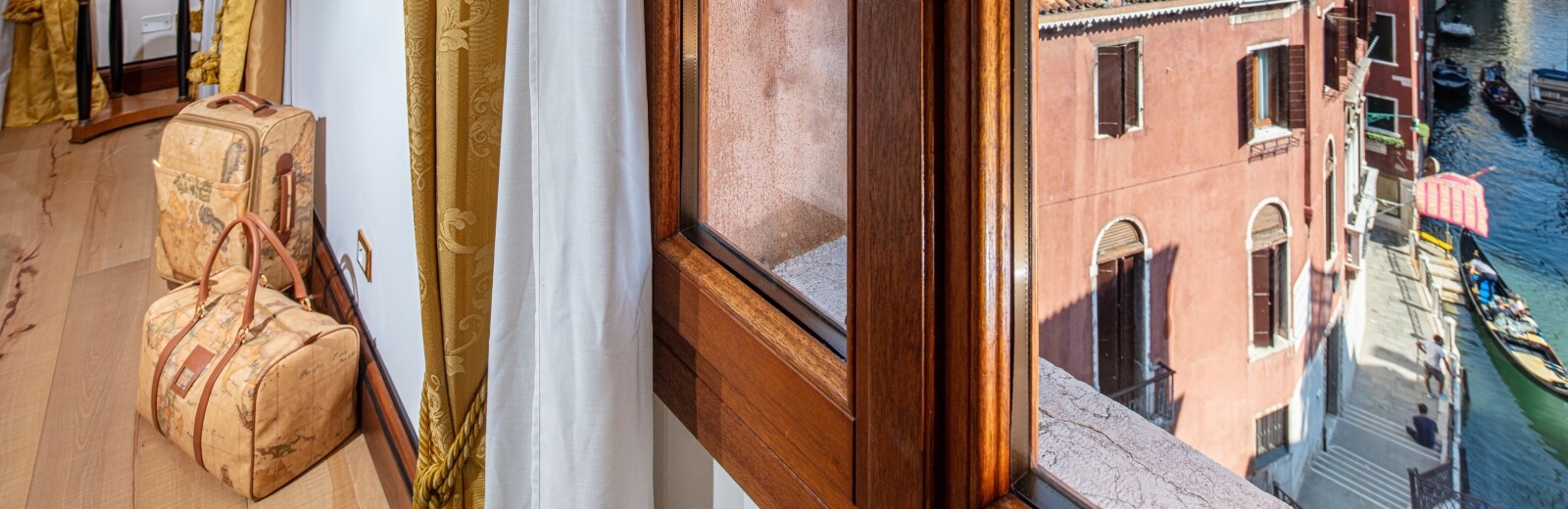 Vista di una camera d'hotel veneziana con luce naturale, valigie eleganti e un canale pittoresco con barche.