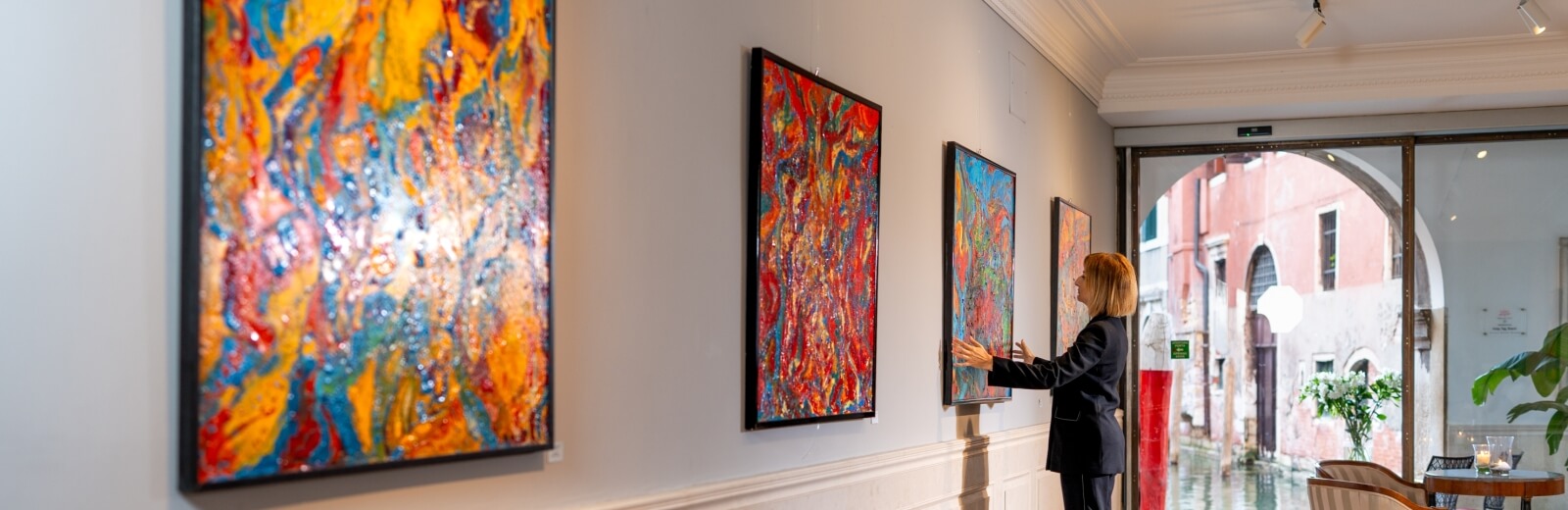 Donna bionda in giacca scura osserva un dipinto astratto colorato in una galleria d'arte con vista su un canale di Venezia.