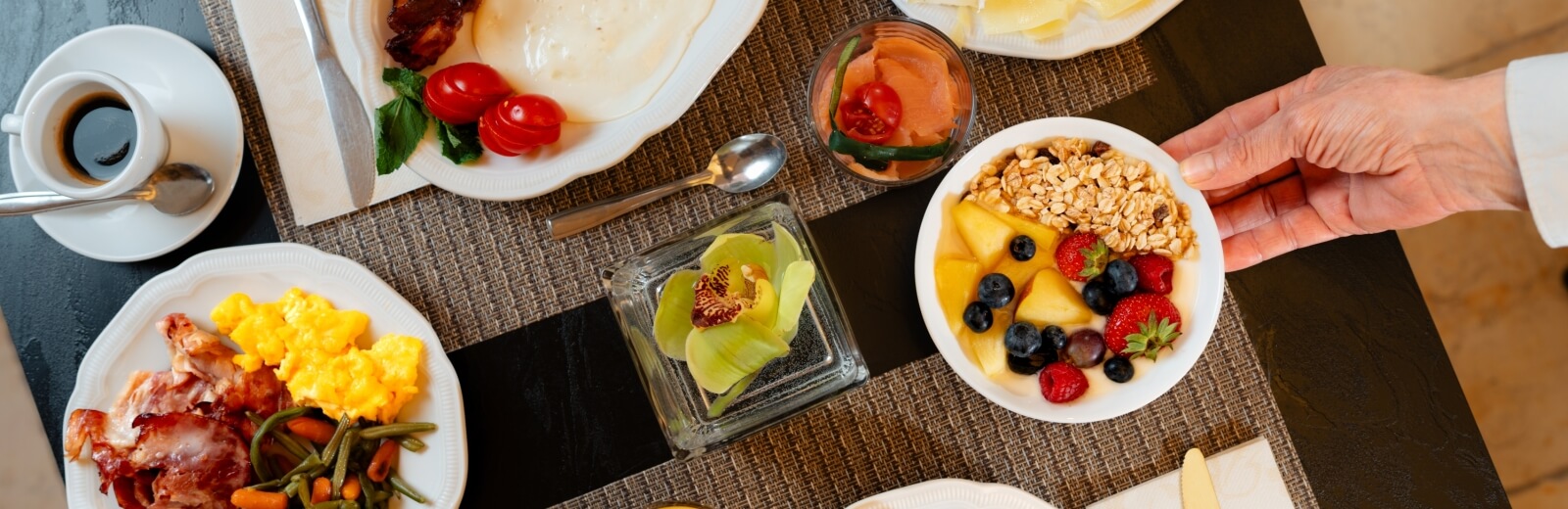 Tavolo con cibo: caffè, bacon, uova, verdure, salmone, yogurt con frutta, mano che tiene una ciotola.