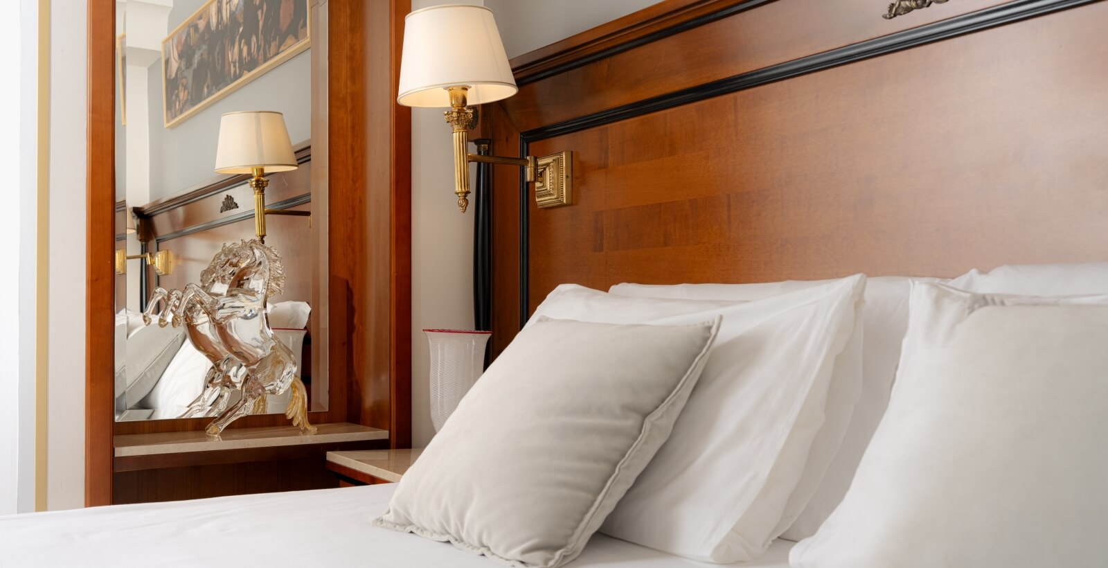 Camera da letto serena con letto bianco, testiera in legno, applique dorata, specchio e scultura di cavallo in vetro.