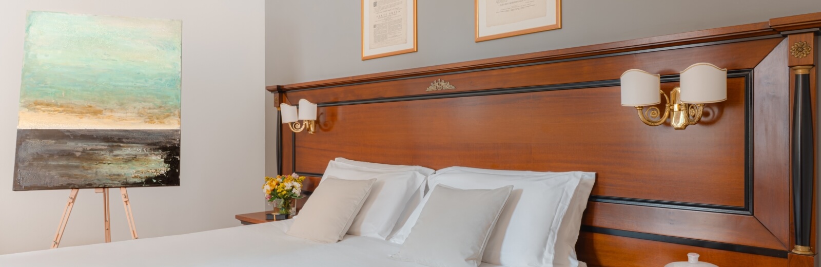 Camera da letto serena con letto elegante, testata in legno scuro, lampade bianche e un dipinto colorato su un cavalletto.