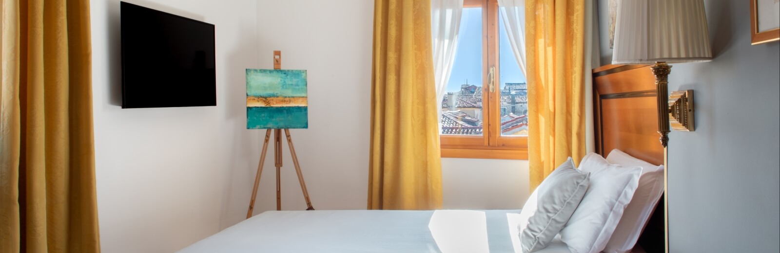Camera luminosa con letto bianco, finestra con tende gialle e un dipinto su un cavalletto a sinistra.
