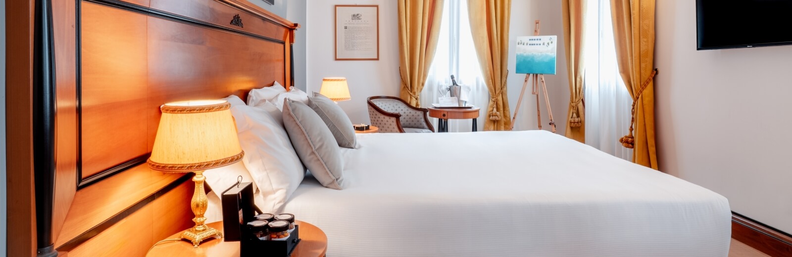 Camera d'albergo elegante con letto grande, tende gialle, lampade e un dipinto su un cavalletto, atmosfera confortevole.