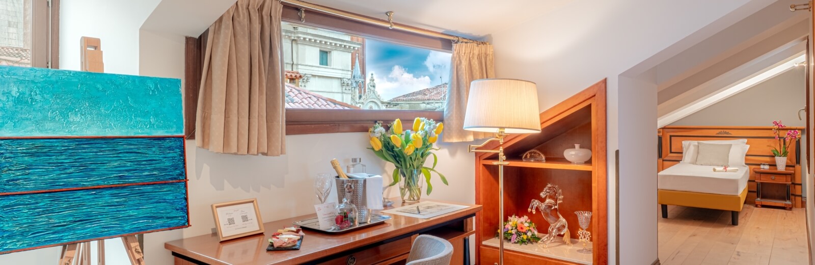 Interior of a cozy hotel room featuring a blue painting, desk with yellow tulips, and a view of a white building outside.