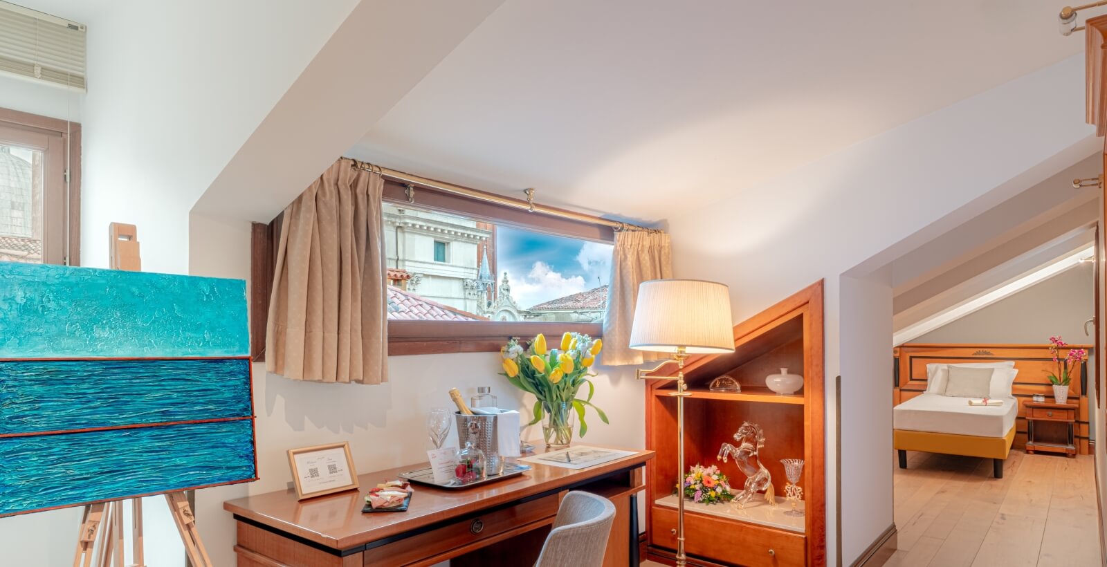 Una stanza ben illuminata con un desk, lampada, decorazioni, un dipinto e una porta che conduce a una camera da letto.