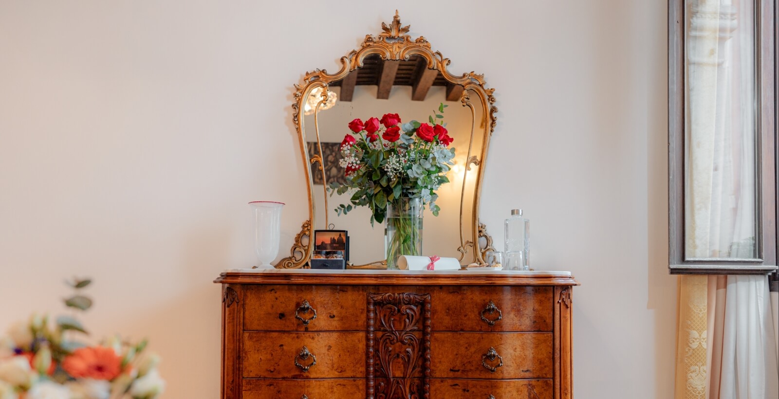 Comò in legno scuro con specchio dorato, fiori rossi e decorazioni, su sfondo bianco con finestra e tende beige.