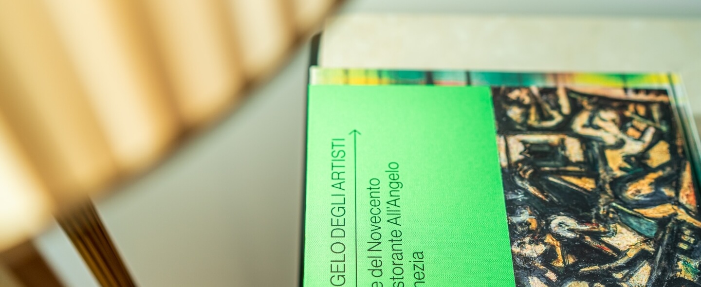 Libro verde con arte astratta in copertina, titolo parzialmente visibile: "GELO DEGLI ARTISTI". Lampada dorata sfocata sullo sfondo.