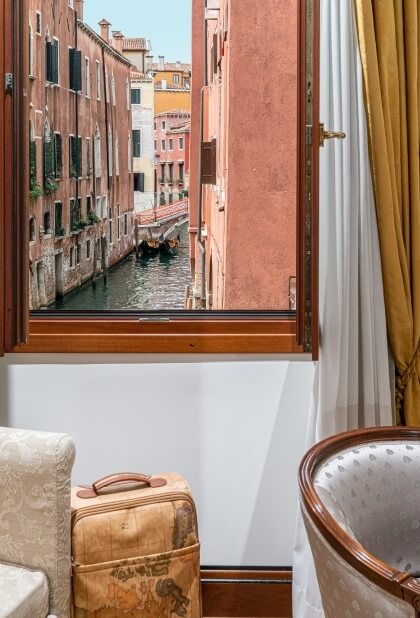 Finestra aperta su un canale di Venezia, con vista su acqua calma e un piccolo battello, in una stanza tradizionale.
