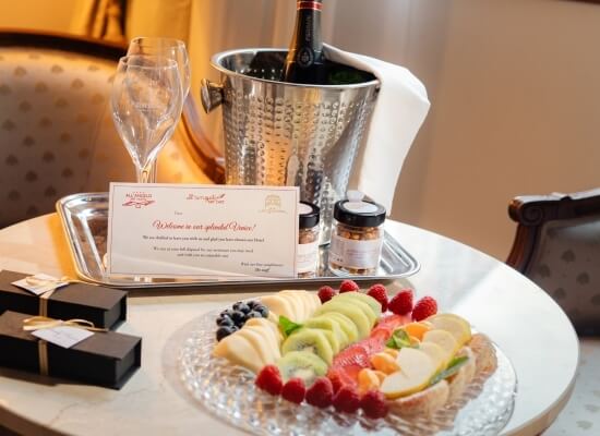 Impostazione di un tavolo lussuoso con vino, frutta e un biglietto di benvenuto in un ambiente di hotel elegante.