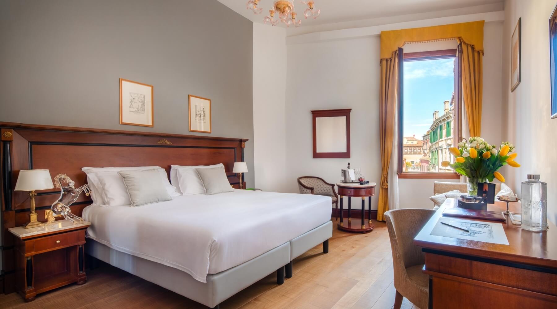 Camera d'hotel elegante con letto, scrivania, sedia e vista sulla strada attraverso una finestra con tende gialle.