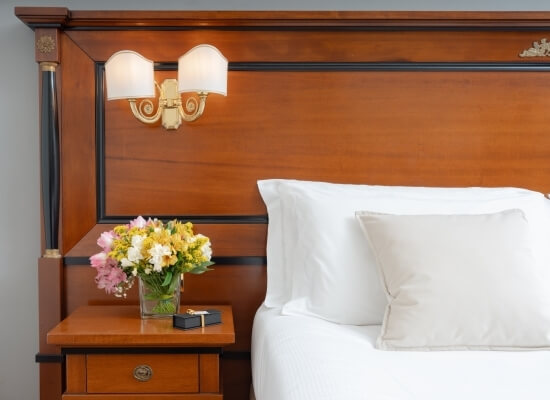 Camera d'hotel lussuosa con letto elegante, biancheria bianca, bouquet di fiori e un comodino raffinato.