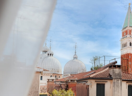 Linee di tetto con torri e edifici a tegole rosse, sotto un cielo blu, con un velo bianco che sfoca l'immagine.
