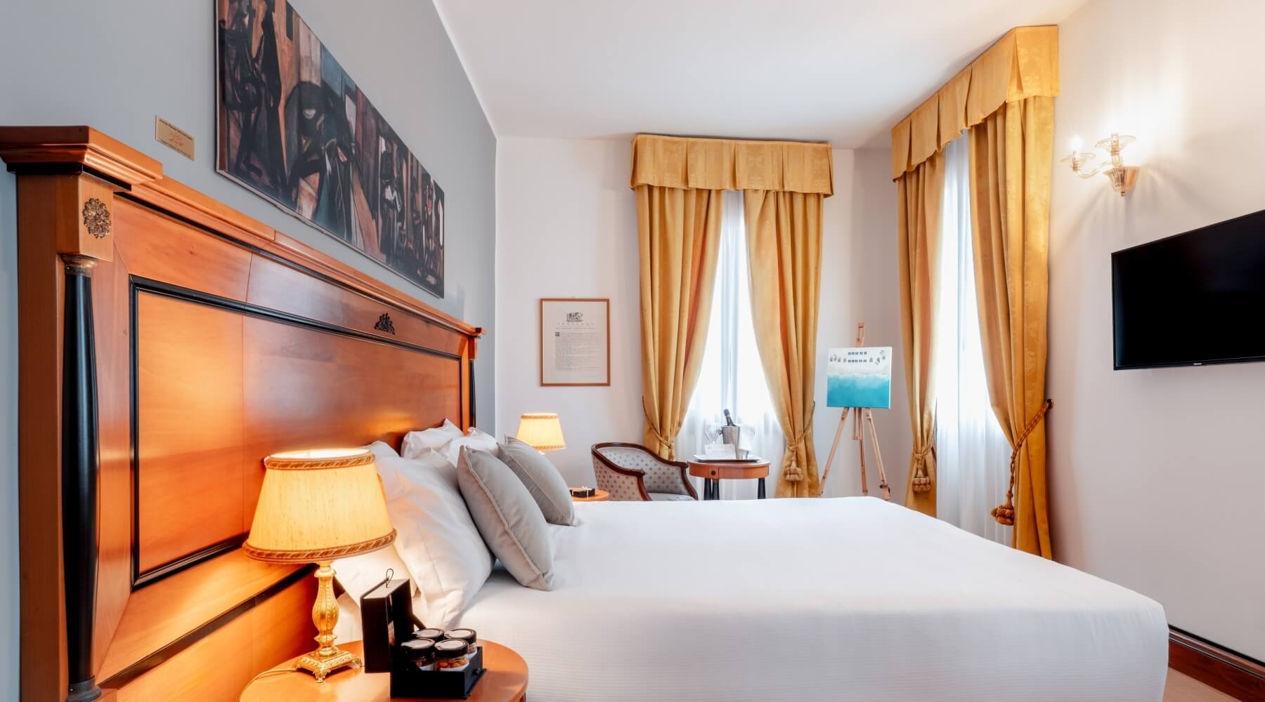 Camera d'albergo accogliente con letto grande, comodino, opere d'arte e finestre con tende gialle.