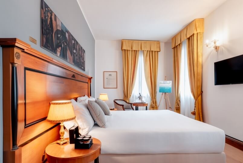 Camera d'hotel accogliente con letto grande, testiera in legno, lampada, TV e finestre con tende gialle.