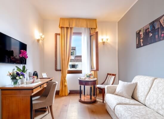 Soggiorno accogliente con divano bianco, scrivania in legno, fiori, pittura e TV, illuminato da una grande finestra.