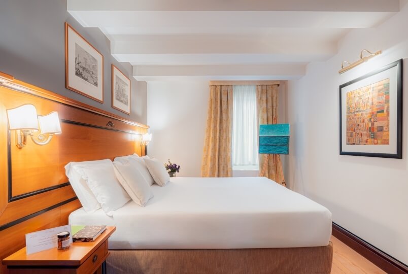 Camera serena con letto bianco, comodino, sconce illuminate, finestra con tende leggere e opere d'arte. Atmosfera tranquilla.