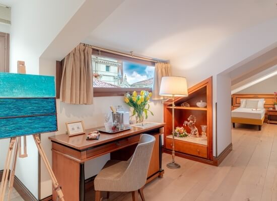 Una stanza con scrivania in legno, sedia, scaffali decorativi, letto bianco e un cavalletto con un dipinto di acqua.