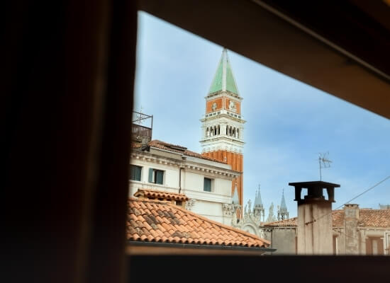 Cityscape viewed through an open window, featuring a tower with orange stripes, red-tiled roofs, and a blue sky.