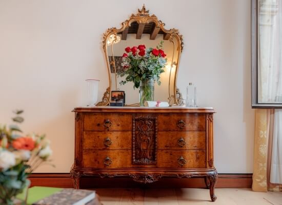 Comò in legno con specchio, fiori, cornice e bottiglie, riflesso del soffitto, finestra con tende bianche sullo sfondo.