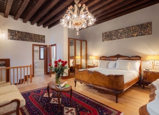 Camera elegante con letto ornato, lampade, vasi di rose e un lampadario, su un pavimento in legno chiaro.