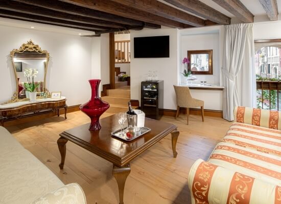 Camera spaziosa in stile veneziano con mobili eleganti, letto a strisce pesca, tavolino e balcone con tende bianche.