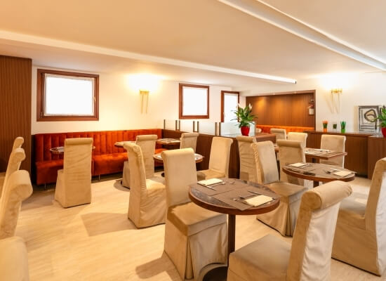 Ristorante elegante con pareti bianche, pavimenti in legno chiaro, tavoli rotondi e booth rossi. Piante decorative presenti.