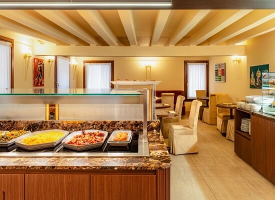 Stanza spaziosa con buffet di cibo a sinistra e area pranzo a destra, illuminata da luce naturale e decorata con opere d'arte.