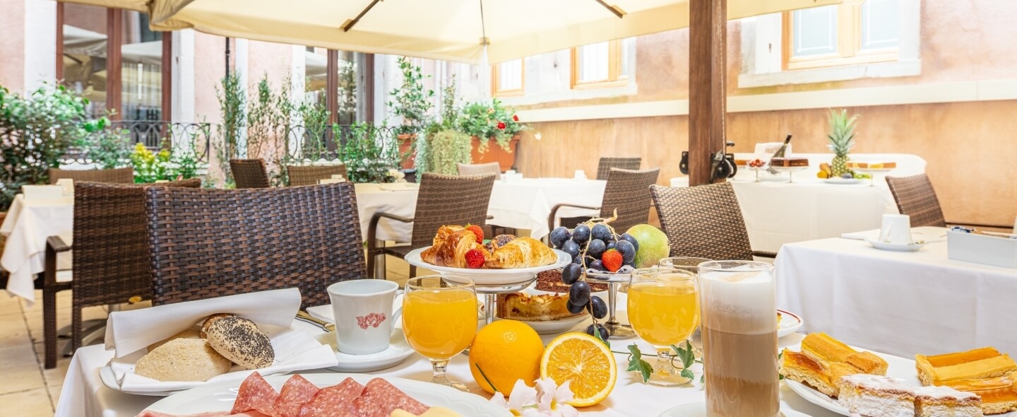 Buffet di colazione con pane, affettati, caffè, succo d'arancia, croissant, frutta e una torta su tavolo bianco.